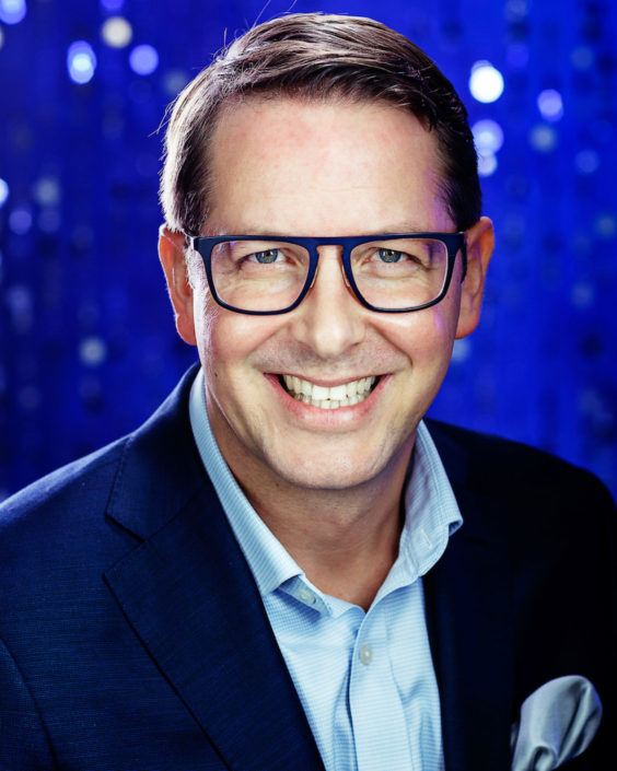 Middle aged smiling businessman wearing glasses blue suit no tie but hanky in pocket with electric blue background