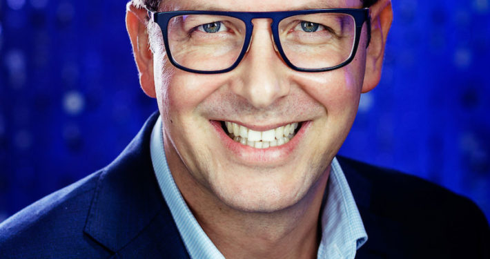 Middle aged smiling businessman wearing glasses blue suit no tie but hanky in pocket with electric blue background