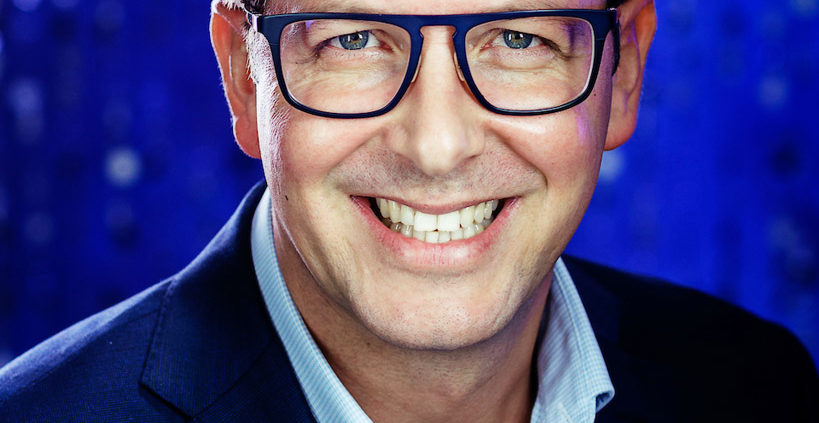 Middle aged smiling businessman wearing glasses blue suit no tie but hanky in pocket with electric blue background