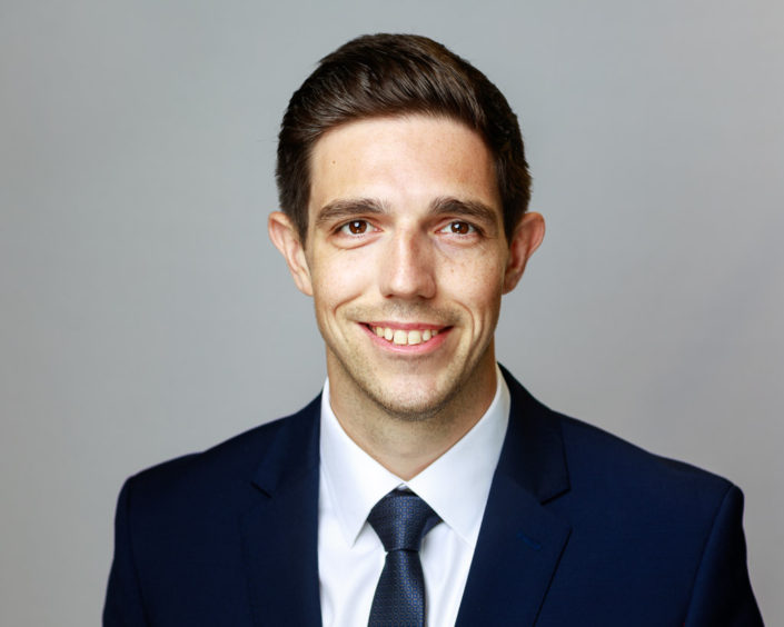 Young business man in blue suit with white shirt smiling at the camera wearing a tie and light grey background behind him