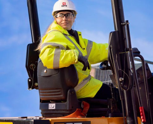 A female construction worker in PPE reversing mining digger with blue sky behind