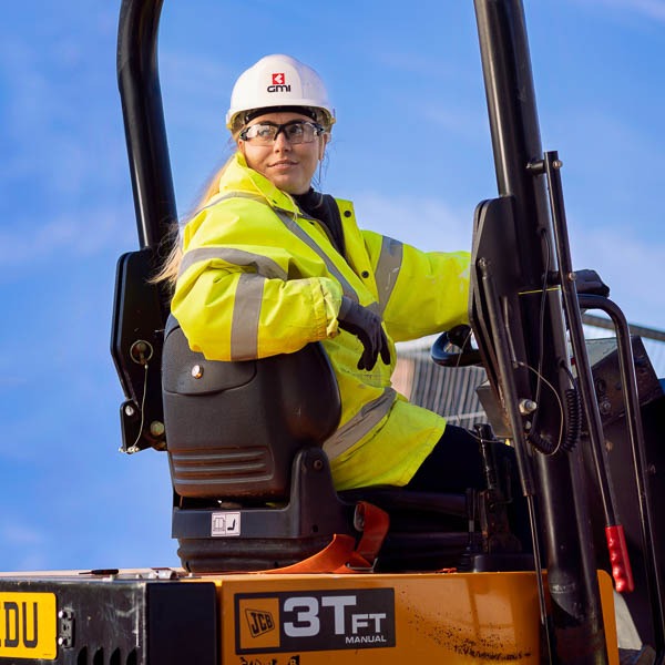 A female construction worker in PPE reversing mining digger with blue sky behind