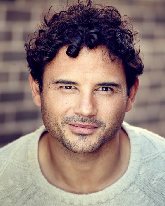 Actor headshot portrait featuring Ryan Thomas taken outside against brown brick wall with Ryan smiling